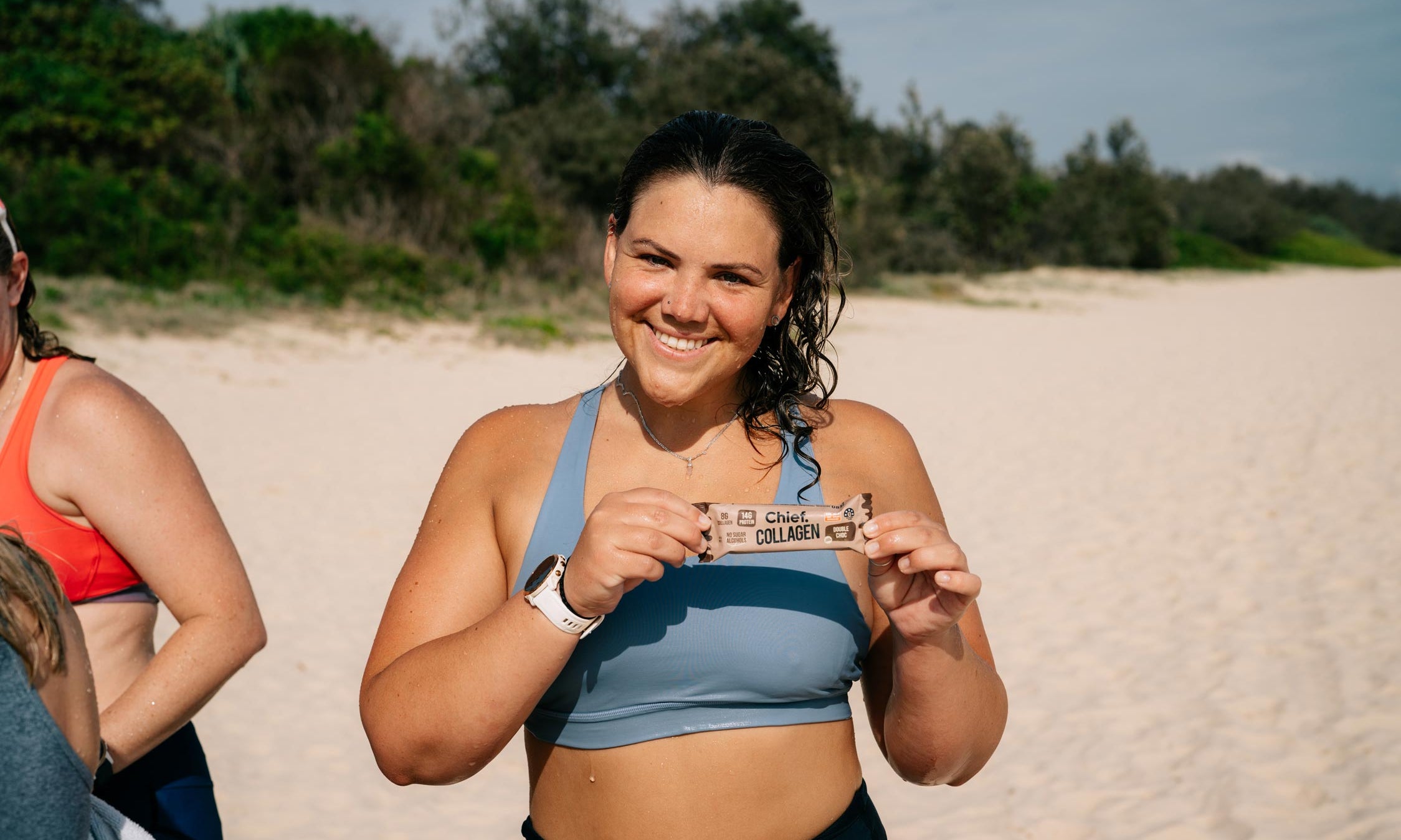 A woman on the beach in activewear smiling at the camera and holding up a Chief Collagen Bar