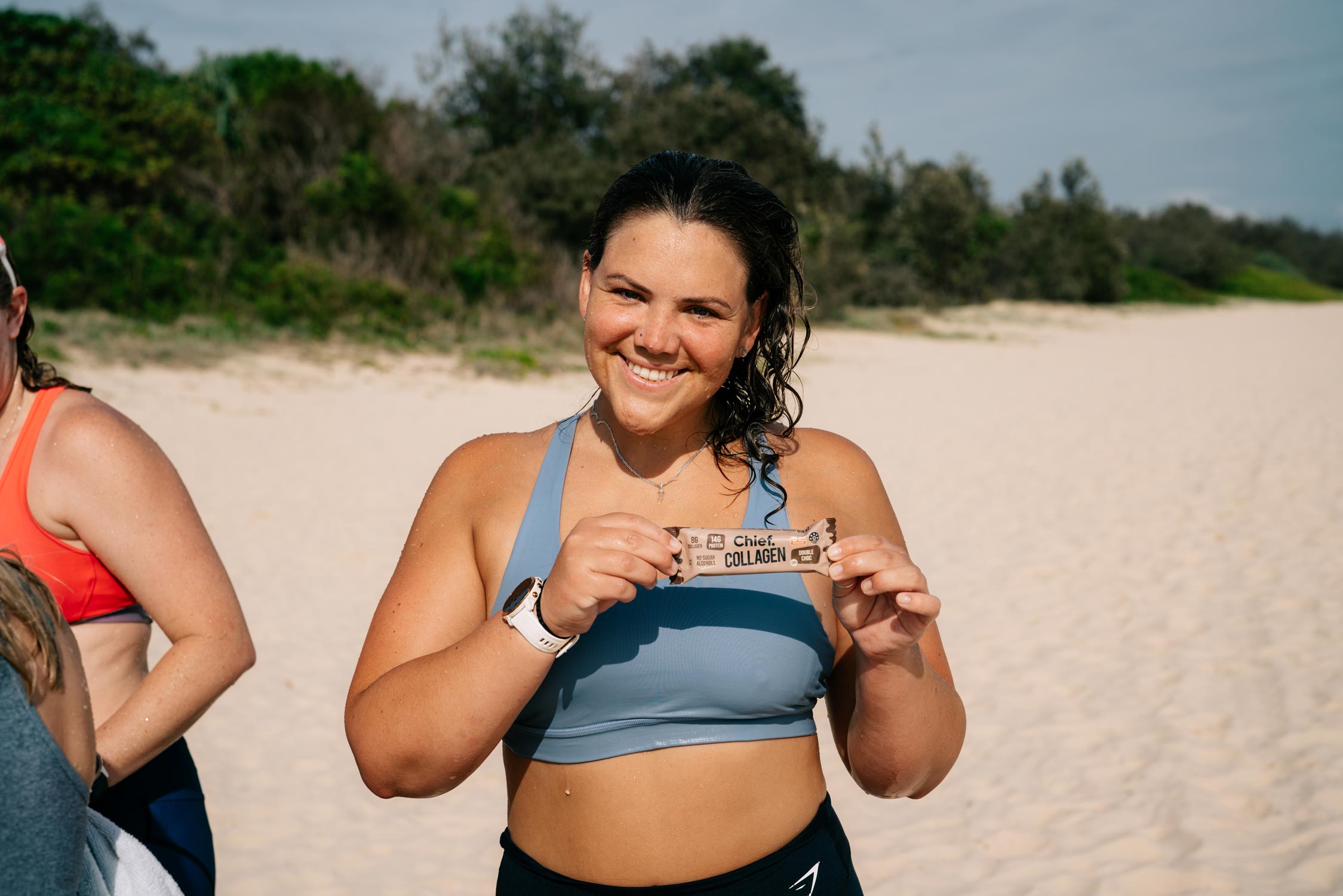 A woman on the beach in activewear smiling at the camera and holding up a Chief Collagen Bar