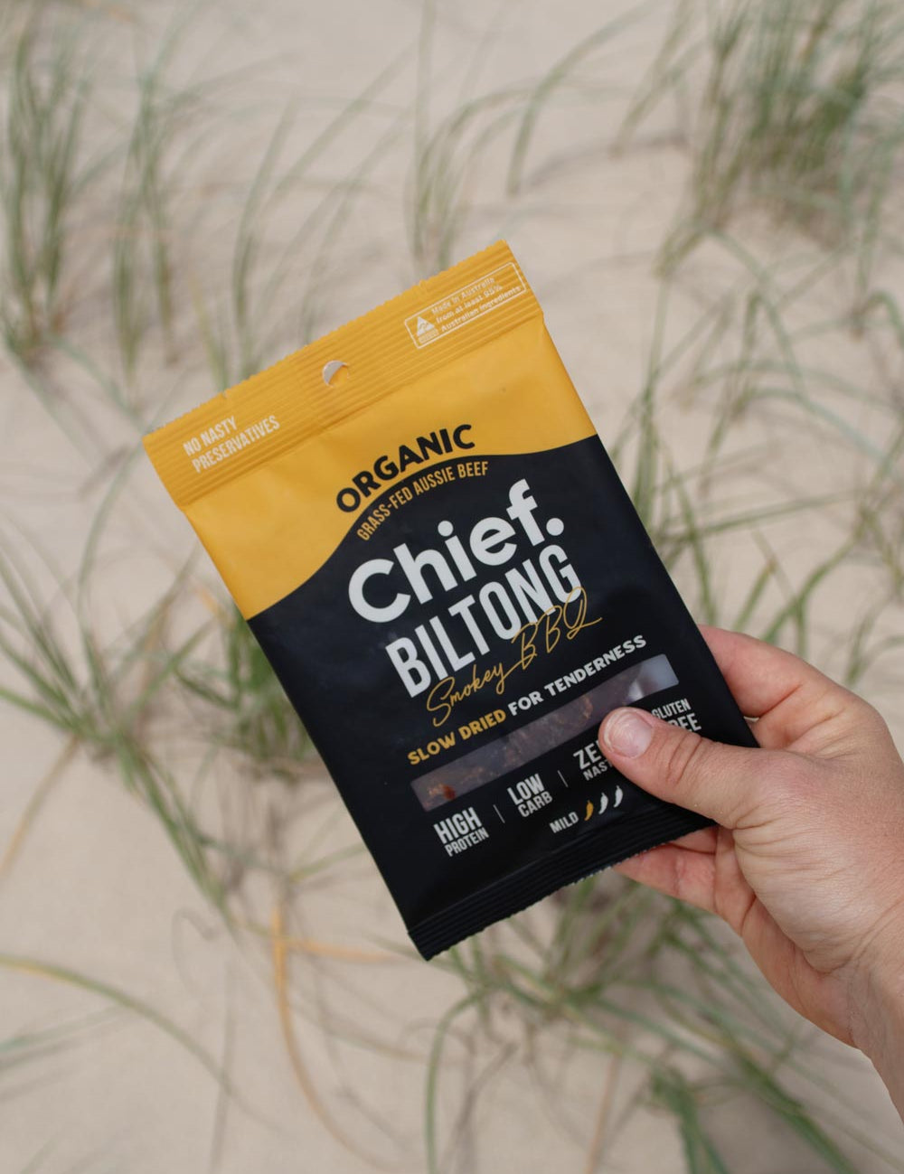 A person's hand holding a bag of Chief Smokey BBQ Biltong against a sand dune backdrop