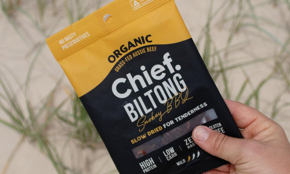 A person's hand holding a bag of Chief Smokey BBQ Biltong against a sand dune backdrop